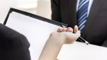 Two people in a meeting, one holding a clipboard with documents.